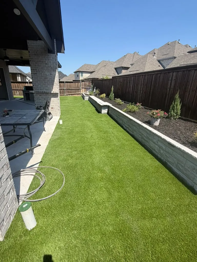 Artificial turf yard beside a raised stone planter and covered patio